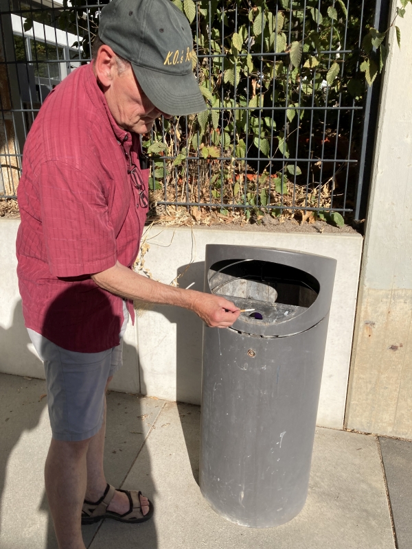 Heinrich Schlosser demonstriert, wie man seine Zigaretten-Kippe nach dem Ausdrücken am Deckelrand des Mülleimers in der kleinen Öffnung ordentlich entsorgt. Dieser „Aschenbecher“ ist im Inneren getrennt vom Rest des Mülls. Foto: Weinmann-Abel
